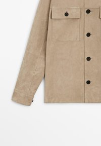 Beige Wildlederjacke mit schwarzen Knöpfen, Klappbrusttaschen und langen Ärmeln vor weißem Hintergrund.