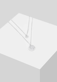 Collane a strati in argento con pendenti circolari dalle intricate decorazioni floreali traforate, esposte su una piattaforma geometrica bianca.