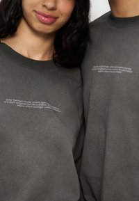 Δύο άτομα φορούν σκούρα γκρι πουκάμισα με μια κυρτή λευκή κείμενη επιγραφή κοντά στο στήθος που αναγράφει μια εμψυχωτική φράση για την πτήση και το θάρρος.