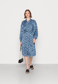 Vestido azul claro com padrão de estampa de leopardo, frente com botões, mangas longas e cinto na cintura, combinado com sandálias escorregadias pretas e uma bolsa preta.