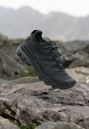 Chaussure de randonnée noire La Sportiva en l'air au-dessus d'un terrain rocheux avec un arrière-plan montagneux flou.