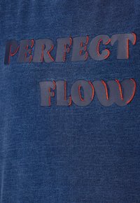 Tessuto blu navy con testo ricamato in rilievo "PERFECT FLOW" di colore rosso in contrasto, con una consistenza liscia e un design casual.