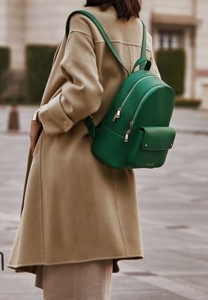 Personne portant un manteau beige et un sac à dos en cuir vert avec plusieurs compartiments zippés dans un environnement urbain en plein air.