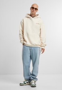 Beige kapucnis felső, elülső zsebbel és logó részlettel, világos kék farmernadrággal és zöld színű kiegészítőkkel ellátott sportcipővel párosítva. Sima textúra, laza szabás.