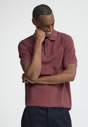 Smilende mand iført en bordeaux polo og mørke bukser, hviler hagen på hånden med armene krydset mod en ensfarvet baggrund.