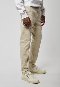 Pantaloni beige in cotone misto con taglio dritto, caratterizzati da una trama sottile e una chiusura con zip. Abbinati a sneakers bianche in pelle.