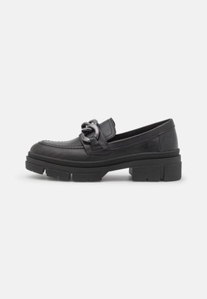 Mocassin chunky noir avec semelle épaisse antidérapante et grand détail chaîne sur le dessus, vu de profil sur fond blanc.