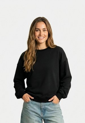 Jeune femme aux longs cheveux ondulés portant un sweat-shirt noir et un jean bleu clair, souriante, les mains dans les poches, debout devant un fond blanc.