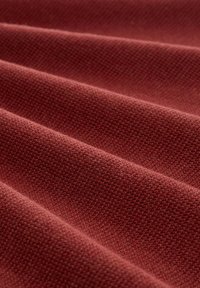 Tessuto con texture in una tonalità di rosso profondo, caratterizzato da un motivo intrecciato. La superficie è morbida, cade in pieghe delicate.