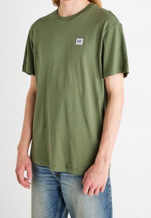 Personne portant un t-shirt vert olive à manches courtes avec un petit patch logo Lee sur la poitrine et un jean en denim bleu clair.