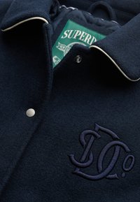 Chaqueta de lana azul marino con un acabado texturizado, que presenta un botón de presión plateado, ribete blanco y un logotipo bordado en la parte delantera. Etiqueta de la marca en verde visible.