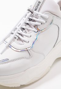 Zapatilla de cuero blanca con acentos plateados, que presenta un detalle reflectante multicolor en el costado y cordones blancos. Suela blanca gruesa.