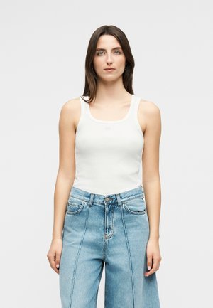 Jeune femme aux longs cheveux bruns portant un haut blanc sans manches et un jean bleu taille haute, debout devant un fond clair uni.