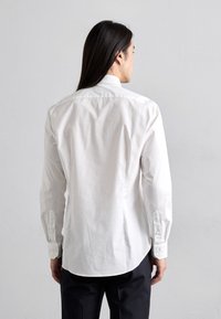 Persona con lunghi capelli scuri che indossa una camicia bianca a maniche lunghe e pantaloni neri, rivolta verso una parete grigia chiara e liscia.