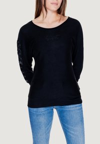 Maglietta nera a maniche lunghe con scollo ampio, caratterizzata da un tessuto testurizzato e lettering in rilievo lungo la manica sinistra. Abbinata a jeans blu.