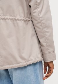 Veste beige clair avec une texture lisse, dotée d'une taille cintrée, de manches longues et d'un poignet replié, assortie à un denim bleu clair.