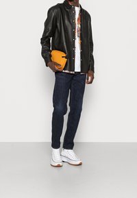 Giacca di pelle nera sopra una camicia bianca con fantasia multicolore, jeans blu scuro, borsa clutch gialla e sneakers bianche con suola in gomma.