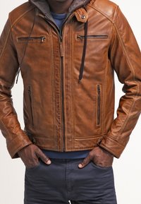 Veste en cuir marron avec capuche, dotée de poches zippées et de détails de couture visibles. Texture douce et coupe ajustée, conçue pour la chaleur.