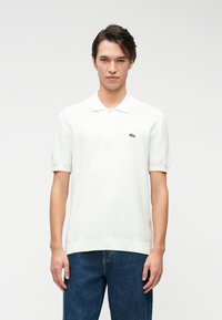 Polo shirt blanc avec un design en tricot texturé, manches courtes, et un logo brodé vert sur la poitrine gauche. Porté avec un jean en denim bleu.