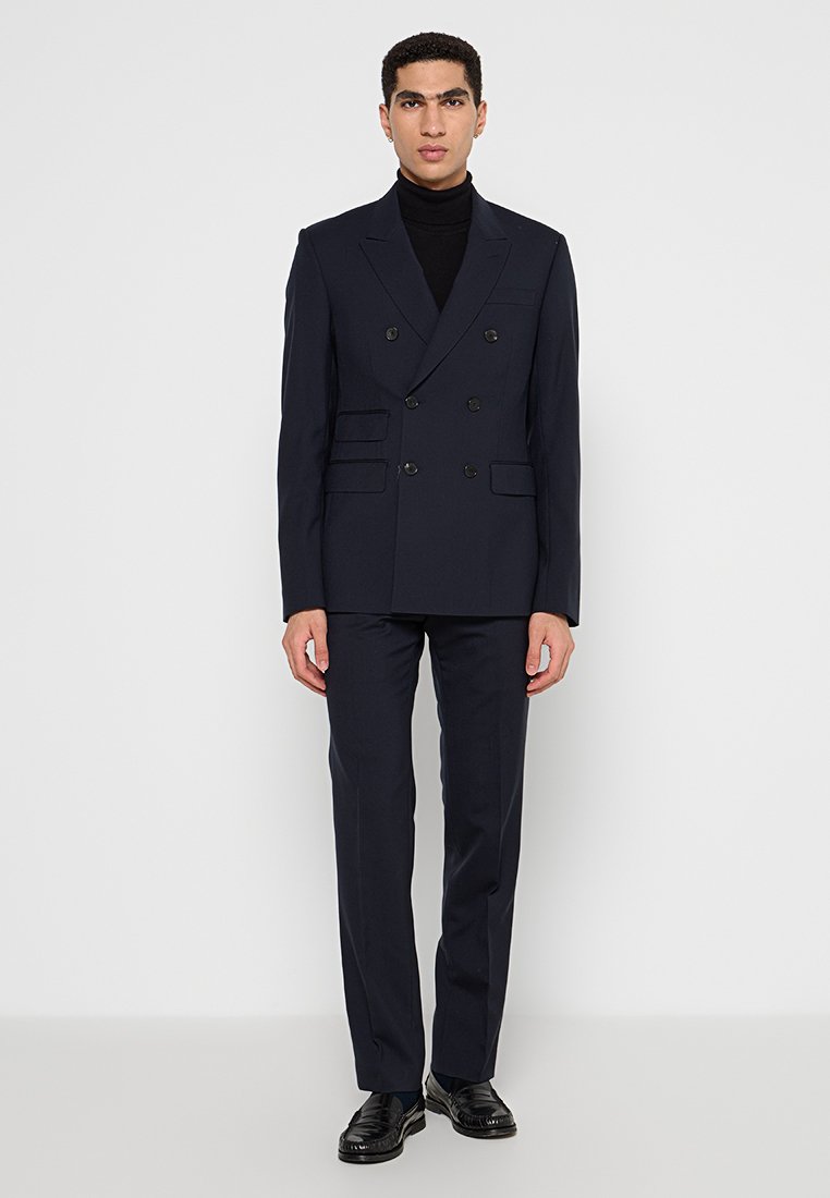 Chaqueta de traje azul marino de doble botonadura con hombros estructurados, tres bolsillos delanteros y pantalones entallados a juego. Acompañada por un cuello de tortuga negro.