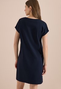 Femme aux longs cheveux portant une robe bleu marine à manches courtes, debout de dos sur un fond beige uni.