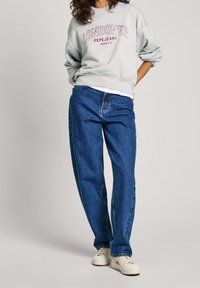 Sudadera gris claro con el texto "LONDONER", combinada con jeans de mezclilla oscuros y sueltos y zapatillas blancas, mostrando un diseño de atuendo casual.