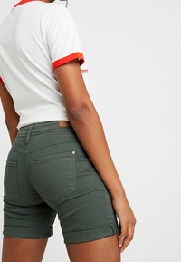 Camiseta blanca con detalles rojos combinada con pantalones cortos de mezclilla verde. Los pantalones cortos tienen dobladillos enrollados y detalles en los bolsillos, con una etiqueta de marca de cuero marrón.