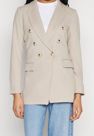 Kobieta ubrana w beżową dwurzędową marynarkę z złotymi guzikami, białą koszulę pod spodem i niebieskie dżinsy, widoczna od ramion do bioder.