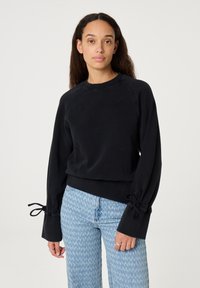 Zwarte cropped sweatshirt met lange mouwen en strikdetails bij de manchetten, gemaakt van zachte stof. Gecombineerd met lichtblauwe gemusterde jeans.