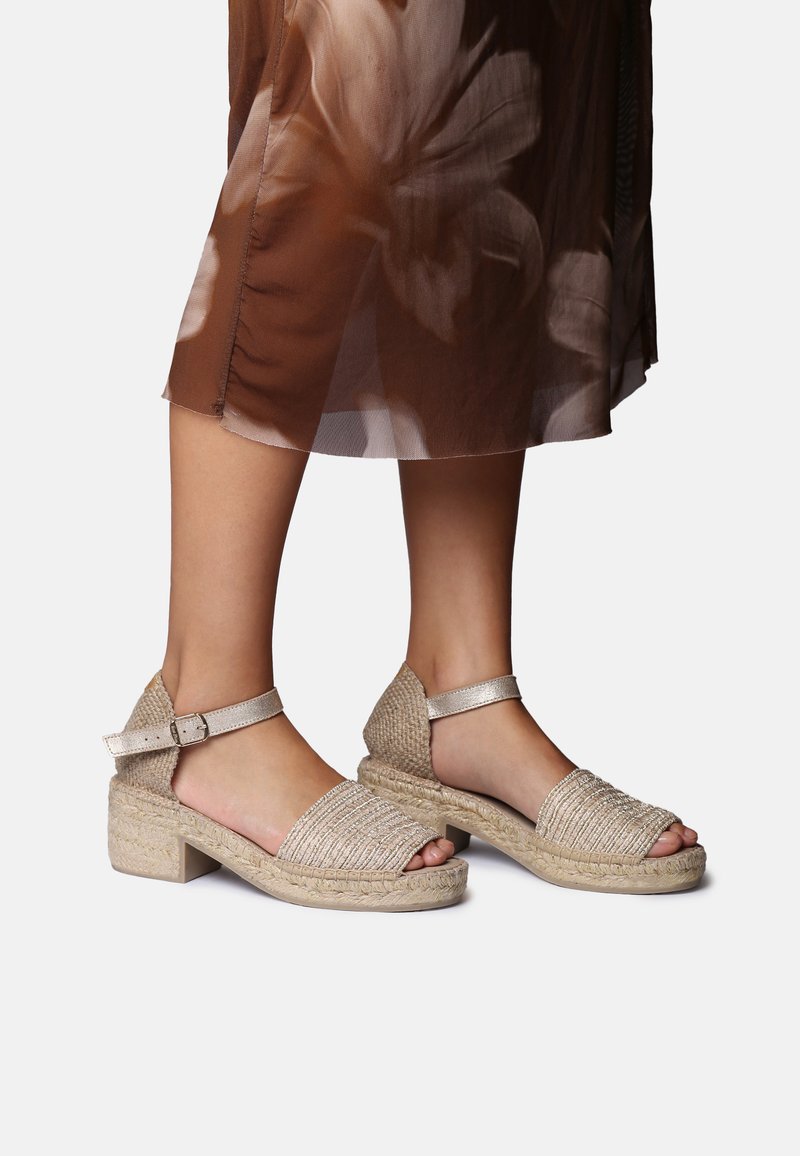 Fødder iført beige flettede sandaler med ankelrem og blokhæle, kombineret med et flydende brunt skørt med et lyst blomsterprint.