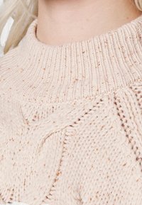 Gros plan sur un pull en laine torsadée beige clair avec un col montant côtelé porté par une personne à la peau claire et aux cheveux blonds.