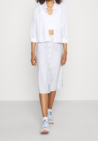 White cropped shirt over a white tank top, paired with a knee-length striped skirt featuring buttons. Light sneakers with pastel accents.