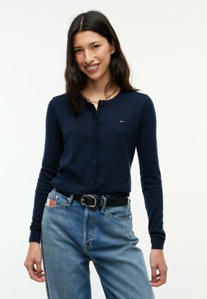 Junge Frau lächelt, trägt eine marineblaue, geknöpfte Strickjacke, hellblaue Jeans mit schwarzem Gürtel und eine goldene Halskette.
