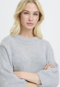 Femme blonde aux yeux bleus portant un pull en laine gris clair, se tenant les bras croisés sur un fond blanc uni.