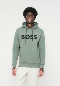 Sudadera verde con un bolsillo frontal, logo "BOSS" en negro, capucha con cordón y puños de canalé. Modelo con pantalones de color claro.