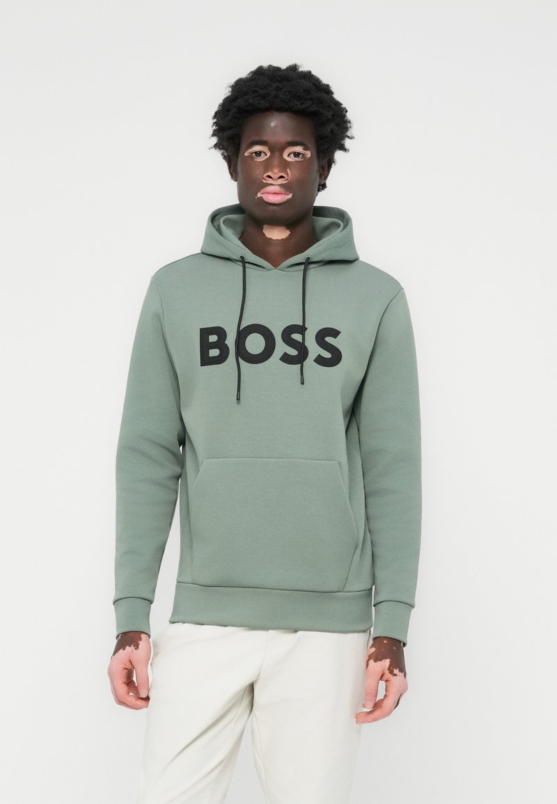 Sudadera verde con un bolsillo frontal, logo "BOSS" en negro, capucha con cordón y puños de canalé. Modelo con pantalones de color claro.