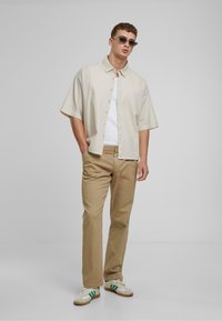 Cămașă scurtă bej deschis cu mâneci scurte, maieu alb dedesubt, pantaloni bej, ochelari de soare gri și adidași cu accente verzi.