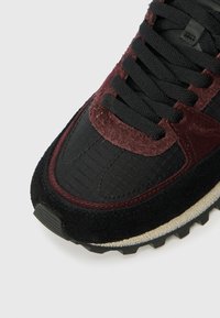 Close-up of a black and dark red sneaker toe with textured suede, black laces, and a rugged white and black sole.