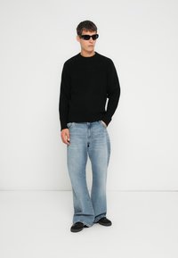 Zwarte gebreide trui met een ronde hals, gecombineerd met lichtblauwe wijde jeans. Matte zonnebril maakt de outfit compleet.
