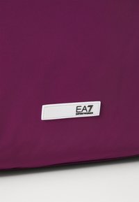 Lilla stoffoverflate med et lite hvitt rektangulært merke som viser "EA7 EMPORIO ARMANI"-logoen i svart tekst.
