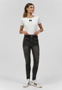 Haut blanc à manches courtes avec des bordures noires et un détail nœud, associé à un jean skinny noir taille haute et des baskets blanches avec des rayures noires.