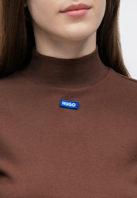 Pruun soonikkoega kõrge kaelusega pluus, millel on rinnal sinise tikandiga "HUGO" silt, tihedalt istuv lõige.