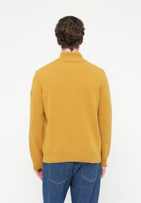 Mustársárga kötött pulóver magas nyakkal, hosszú ujjakkal és bordázott szegéllyel. Az anyag puhanak tűnik, finom textúrával.
