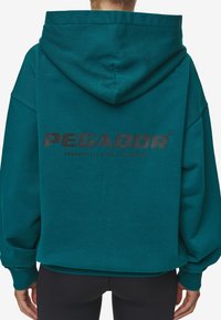 Felpa teal con vestibilità rilassata, grande cappuccio e polsini a costine. Presenta un logo nero sulla schiena. Texture morbida, design casual.