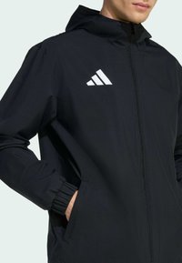 Μαύρο αδιάβροχο μπουφάν με μπροστινό φερμουάρ, ελαστικούς καρπούς και λευκό λογότυπο με τρεις ρίγες στο αριστερό στήθος.