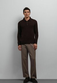 Jonge man die een donkerbruine polotrui met lange mouwen draagt, beige broek met wijde pijpen en zwarte schoenen, staand tegen een effen achtergrond.