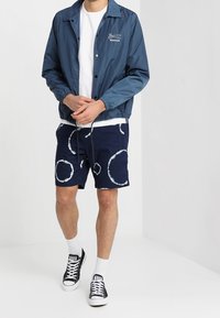 Námořnická bunda se zipem s bílým logem, doplněná námořnickými šortkami s bílými kruhovými vzory. Černé tenisky s bílými detaily.