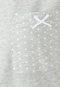 Tessuto grigio chiaro con una texture morbida, caratterizzato da una tasca a pois. La tasca ha pois bianchi e un piccolo accento a forma di fiocco bianco.