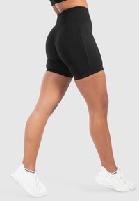 Shorts noirs taille haute en tissu extensible, présentant un design ajusté, une poche latérale et des coutures contrastées. Portés avec des baskets blanches.