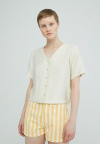 Beige blouse met korte mouwen en v-hals, voorzien van knopen, gecombineerd met gele en witte gestreepte shorts. Zachte stof en ontspannen pasvorm.